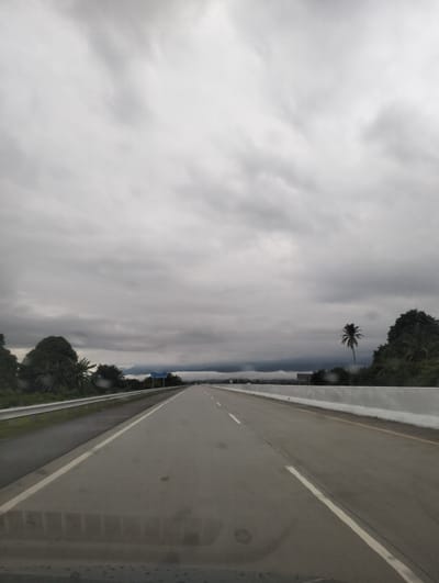 The image depicts a highway stretching into the distance under a cloudy sky. The road is paved and has white lane markings. On the left side of the road, there is a metal guardrail, and beyond that, a line of trees. On the right side of the road, there is a concrete barrier. In the distance, mountains can be seen through a layer of clouds. There is a sign on the left side of the highway with some text, but it is illegible. A lone palm tree stands in the middle ground on the right. The overall atmosphere is overcast and slightly damp, suggesting it might be raining or has recently rained, as evidenced by the droplets on the windshield in the foreground. The location is identified as Baksukon, Indonesia. There are no people or vehicles visible in the image.