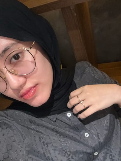 A light-skinned individual is depicted in an indoor, close-up portrait, located in Patrang, Indonesia. The subject, a female, wears a black hijab covering her head and neck, and a dark grey collared shirt with a subtle, repeating patterned texture and white buttons. She wears round eyeglasses with gold-colored frames and a distinct upper bar accent. Her lips are visible and appear glossy with a light pink tint. Her right hand is in the frame, with two gold-toned rings: one with a clear stone on the ring finger and another with a small heart or knot design on the middle finger. The individual gazes directly towards the camera. The background is dimly lit and indistinct, showing dark brown, possibly wooden, architectural elements in the upper right.