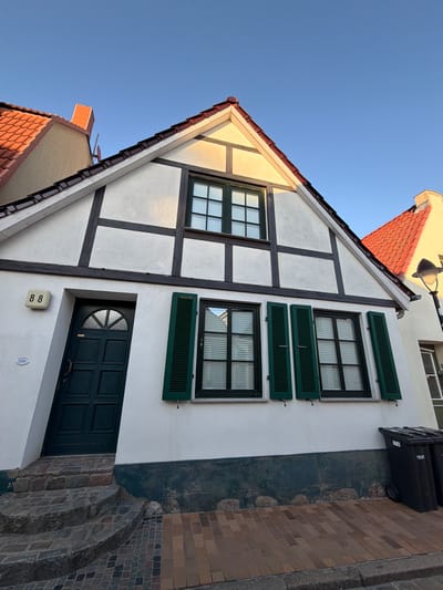 The image shows a close-up, low-angle exterior shot of a historic half-timbered building located in Rostock, Germany. The building features white stucco walls with dark timber framing, a red-tiled roof, and green shutters on its windows. A dark green door with a semicircular glass pane is visible on the left side of the facade, above a set of stone steps. Above the door, the house number "88" is displayed on a white plaque. The street in front of the house is paved with bricks. The sky is clear and blue, indicating fair weather. There are no people or animals visible in the frame. The scene is devoid of any activity, presenting a static architectural view.