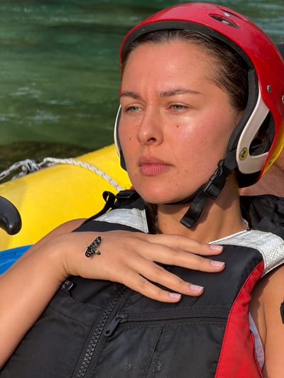 A close-up shot focuses on a young woman wearing a red and yellow helmet and a black and red life vest. She is positioned in what appears to be a yellow raft, with green water visible in the background. A small black and white insect with yellow markings is resting on the back of her left hand, which is placed over her chest. The lighting suggests it is daytime, with the sun highlighting the woman's face and the insect. The overall scene conveys a sense of outdoor activity, possibly on a river or lake.