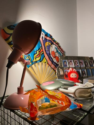An indoor still life scene is depicted, featuring various personal objects arranged on a dark green wire shelving unit against a plain, light-colored wall. Illumination originates from a small, pink, articulated desk lamp positioned on the shelf's left, pointing upwards and to the right, casting a localized glow. A dark electrical cord extends from the lamp.
On the shelf, an open orange plastic bag of "Sour Patch Kids" gummy candies is visible, containing several multicolored gummies, with a single red gummy candy standing upright to its left. To the right of the candy, a silver or grey metallic electronic device, possibly a headphone case, is present, bearing a pink sticker. Adjacent to this, a pair of white Apple AirPods Max headphones are displayed, showing their mesh headband and earcups.
Behind these items, a natural wood-colored folding fan is partially visible. Behind it, a larger, unfolded hand fan with a vibrant graphic design featuring cherries, a crescent moon, and abstract patterns is positioned. To the right, a gold-toned, school bus-shaped photo frame, labeled "SCHOOL BUS," contains multiple small portrait photographs of individuals, likely students. A small, red, plush toy with prominent eyes and ears is situated in front of the photo frame. No living individuals are present.
The precise location within the city of Levallois-Perret, France, cannot be determined from the visual information provided in the image.