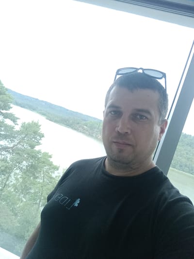 A man is taking a selfie in front of a large window with a view of a lake and a forested hillside. He is wearing a black t-shirt with a light blue logo on the left chest. Sunglasses are perched on top of his head. The man has short, dark hair and a light beard. The window frame, made of grey metal, is visible on the right side of the image. The background shows a body of water, likely Lake Macha, with trees and hills on the far side. The sky is overcast. The man appears to be indoors, possibly in a modern building, given the architecture suggested by the window frame. The overall scene is natural and serene, with the man in the foreground as the primary subject. The location is specified as the city of Doksy, Czechia, which is known for Lake Macha.