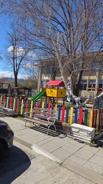 The image depicts a playground situated in Slatino, Bulgaria. The scene includes a colorful play structure with a red roof, yellow walls, and a green slide. The playground is enclosed by a multicolored fence with red, yellow, blue, and green planks. A wooden bench with white metal arms sits adjacent to the playground, alongside a paved walkway. A tree with bare branches is prominently positioned near the playground, partially obscuring the building in the background. A portion of a dark-colored car is visible in the lower right corner. The sky is blue with a few clouds.