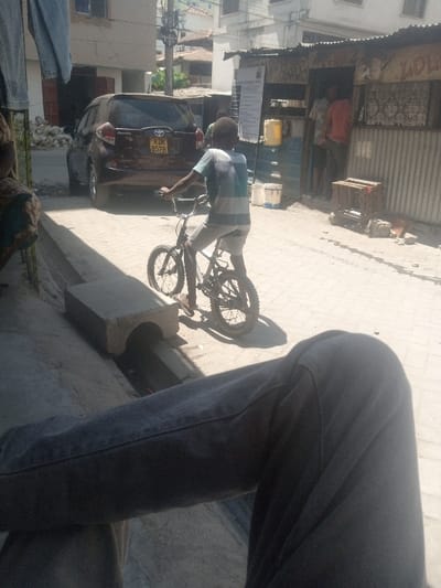 The image depicts a sunny street scene in Tudor ward, Kenya. A young boy with dark skin, wearing a light blue and white striped t-shirt and light-colored shorts, rides a black BMX bicycle towards the right mid-ground. He wears flip-flops. Behind him and to the left, a maroon Toyota SUV with Kenyan license plate KDK 857B is parked on the paved street. To the right, a makeshift structure constructed from corrugated metal sheets serves as a storefront or dwelling, with two partially visible adult males inside. A yellow jerry can and several white buckets are positioned outside this structure, alongside a wooden crate resting on the ground. The street is paved with light-colored interlocking bricks. A concrete drainage channel runs along the left foreground, partially spanned by a rectangular concrete culvert piece. In the immediate foreground, the legs of an individual wearing dark grey trousers are visible, indicating a low observational vantage point. Clothing items are visible hanging on a line in the upper left of the frame. The scene is brightly lit by sunlight, casting distinct shadows.