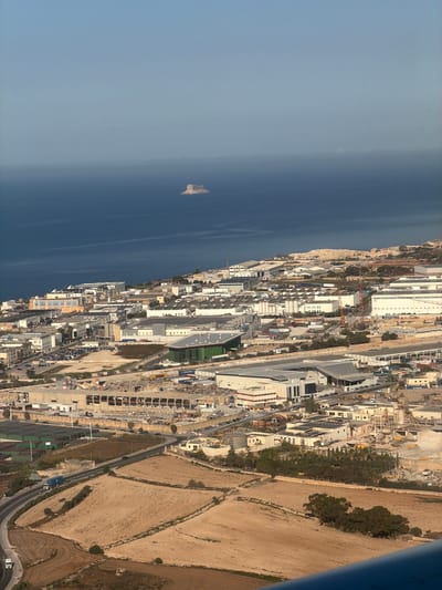This is an aerial, high-angle, long shot photograph of a coastal industrial zone and agricultural fields in the city of Birżebbuġa, Malta, with the Mediterranean Sea and a small island in the background. The sky is a clear, pale blue, with a light haze obscuring the distant horizon. The sea is a deep blue, with a small, rocky island in the middle distance. The foreground consists of agricultural fields with dry, tan-colored soil, interspersed with trees and stone walls. A paved road curves through the fields, with a blue truck and a few cars on it.
The middle ground is dominated by a sprawling industrial complex. Numerous warehouses, factories, and other buildings are visible, many with white or light-colored roofs and walls. Some buildings are modern, with large windows and solar panels, while others appear older and more utilitarian. Several cranes and construction sites suggest ongoing development. Parking lots are filled with cars, and shipping containers are stacked in some areas. A large, green-roofed building stands out among the others.
In the background, the industrial zone continues to extend towards the coast, where it meets the sea. The rocky terrain of the coastline is visible, with further industrial structures built upon it. The small island in the sea is a prominent feature, appearing as a light-colored, rough-textured rock formation. The overall impression is one of a busy, developing industrial area situated along a scenic coastline. There are no people clearly visible in the frame.