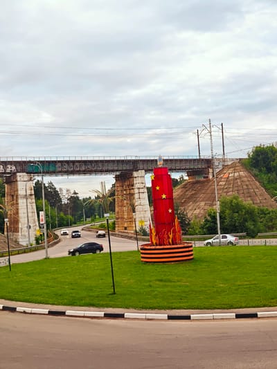 This image depicts a roundabout in the city of Khotkovo, Russia, featuring a large red monument decorated with yellow stars and flames. The monument is situated on a grassy island within the roundabout. In the background, a railway bridge with graffiti spans across the scene, supported by large concrete pillars. Several cars are visible on the road, navigating the roundabout and passing under the bridge. Lush green trees and vegetation surround the area, suggesting a park-like setting. The sky is overcast, with clouds obscuring the sun.