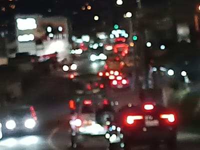 A blurry night scene captures a congested street with various vehicles. Car headlights and taillights create bright, out-of-focus orbs of white and red. The city backdrop features blurred buildings with lit-up signs, contributing to the overall bokeh effect. The image is heavily saturated with light against the darkness, making it challenging to discern specific details beyond the lights themselves.