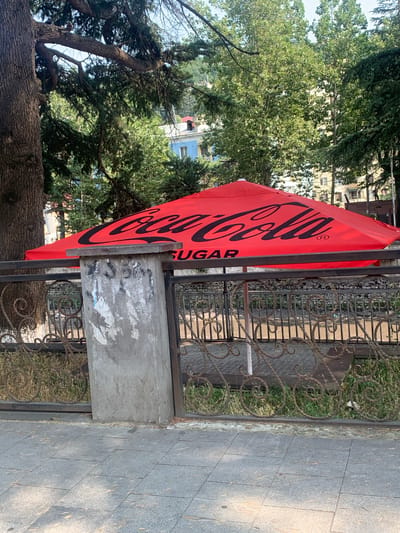 An outdoor scene in Chiatura, Georgia features a large, red Coca-Cola umbrella with the brand's logo prominently displayed in black. The umbrella is supported by a white pole and is positioned behind a decorative metal fence. Adjacent to the fence is a grey concrete pillar with peeling posters on its surface. The ground in front of the fence is paved with square grey tiles, and beyond the fence, there is grass and the suggestion of a river or stream. In the background, lush green trees and some buildings, one painted blue, are visible. The overall impression is that of an outdoor seating area or a public space advertising the Coca-Cola brand.