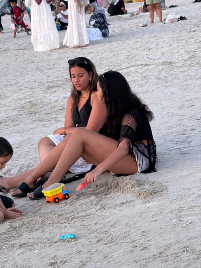 This image depicts a sandy beach scene at Cenang Beach, Malaysia. In the foreground, two adult females are seated directly on the light-colored sand. The female on the left wears a black halter-neck top, white bottoms, black sunglasses on her head, and gold-colored jewelry, including a cross earring and necklace. She has black sandals on her feet. The female on the right wears a black mesh or fringed cover-up over white shorts and holds a pink plastic sand shovel in her right hand, pointed toward the sand. A young male child is partially visible on the bottom left, seated and interacting with the sand. Between these foreground subjects, a small toy dump truck with an orange chassis, blue cab, and yellow bed containing sand is present, along with a small blue plastic toy. In the blurred background, numerous other individuals are visible walking, standing, or sitting on the sand, including a child in a red Spider-Man shirt and adults dressed in various beachwear, some in long white garments or with head coverings. The environment consists solely of sand; no water is visible within the frame.