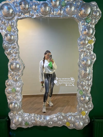 An indoor scene in Dubki (Gp Kubinka), Russia, captured as a mirror selfie. A single female subject stands centered in the reflection of a large, irregularly shaped wall-mounted mirror. She has dark hair styled in braids and wears a white long-sleeved top, black and white tie-dye leggings, white socks, and light-colored athletic shoes. She holds a light-colored smartphone in her right hand, angled to take the photo, and a translucent water bottle in her left hand, over which a folded green towel is draped. Small white earbuds are visible. The mirror features a thick, ornate frame heavily embellished with numerous spherical, silver, disco ball-like ornaments of various sizes, interspersed with small artificial slices of lemon and lime. The immediate wall surrounding the mirror is dark green, where a dark green radiator is visible on the lower left. The reflected background shows a plain light-colored wall with light brown wood or laminate flooring. A white textual graphic is visible on the reflected wall, reading in Cyrillic "ТЫ ВСЕГДА ПРЕКРАСНО ВЫГЛЯДИШЬ" (You always look great) and "LIMB FITNESS" with fruit icons.