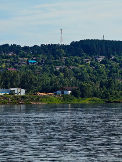 The image depicts a wide, calm river in the foreground, with gentle ripples distorting the reflection of the sky. The riverbank on the opposite side is densely covered with lush green trees, forming a thick forest that climbs up a gentle slope. Nestled within the trees and along the hillside are numerous houses, characteristic of a settlement. Some houses have colorful roofs, such as blue and red.
Further up the slope, two tall communication towers are visible above the treeline, indicating the presence of infrastructure. In the lower part of the image, closer to the riverbank, there are a few buildings that appear to be industrial or storage facilities, with one notable white building with a brown roof and another long, light-colored structure. Piles of logs are also visible on the bank near these structures, suggesting a timber-related activity.
The sky above is a pale blue, with scattered, wispy white clouds. The overall scene conveys a sense of a rural or semi-rural settlement situated along a river, with a backdrop of dense forest and clear skies. The location is identified as the city of Predivinsk, Russia.