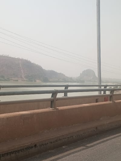 The image displays a panoramic view from a bridge in Jebba, Nigeria, under a hazy sky, likely depicting the River Niger and its surrounding landscape. In the immediate foreground, a dark asphalt road surface is partially visible on the bottom right, with shadows cast upon it. Dominating the lower half of the frame is a reddish-brown concrete bridge barrier, featuring a multi-bar grey metal railing system mounted on its top surface. Beyond the bridge barrier, a wide body of water, appearing calm and light green-grey, stretches across the midground. The far bank of the river is characterized by rolling hills and mountains covered with sparse, faded green vegetation, with visible cleared paths or tracks on some slopes. In the upper mid-ground, multiple parallel power lines or communication cables extend horizontally across the scene. On the far right, a tall, vertical grey pole, possibly a utility or light pole, rises out of frame. In the distant background, obscured by atmospheric haze, further rugged hills and a distinct, prominent rock formation are discernible. No individuals, vehicles, or dynamic actions are present within the frame.