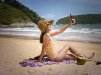 A woman in a yellow and white striped swimsuit and a straw hat sits on a beach in Bevern, Germany. She is holding a smartphone in her right hand, taking a selfie. She is wearing white headphones. A purple towel and a pair of sunglasses are on the sand next to her. A whole coconut with a straw is placed in front of her. The ocean is in the background with waves rolling in. Green hills covered in trees are visible on the horizon. The sky is blue with some clouds.