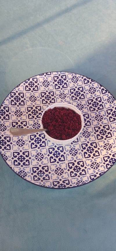 This image shows a close-up, overhead view of a white bowl filled with a reddish-purple granular substance, possibly sumac or a spice blend. The bowl is placed on a decorative ceramic plate with a blue and white Moorish-inspired pattern. A silver spoon rests partially within the bowl, its handle extending downwards. The plate and bowl are situated on a textured, light blue surface, possibly a tablecloth or placemat, with visible lighting and shadow patterns casting across it. The scene appears to be indoors, given the controlled lighting. The focus is on the food item and its presentation. There is no visible text in the image.