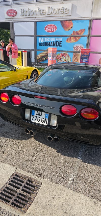 This image captures an outdoor scene in what is presumed to be the city of Bain-de-Bretagne, France. The primary subjects are two sports cars and two women, set against the backdrop of a commercial establishment. In the immediate foreground, a black Chevrolet Corvette is parked, its rear facing the camera. The "CORVETTE" emblem is visible on the trunk lid. It features four distinct circular red taillights, two on each side, and two clear rectangular reverse lights flanking the central license plate. The French license plate reads "F AD-776-GN", with the departmental code "21" (Côte-d'Or) displayed on the right side. Below the bumper, four chrome exhaust tips are visible, arranged in two pairs. The car's surface is glossy, reflecting the surroundings. To the left of the black Corvette, partially obscured, is a bright yellow sports car, likely another Chevrolet Corvette given its body lines and proximity. Only its rear quarter panel, a portion of a wheel, and a side mirror are visible. Two women are standing on the left side of the frame, near the yellow car. The woman closer to the viewer has dark hair and is wearing a pink fuzzy jacket over a dark top and light grey pants. Her mouth is slightly open, and she appears to be holding something to her face or eating. Behind her, an older woman with short, curly grey hair and glasses is visible. She wears a dark jacket with a pink element at the collar. Her left hand rests on a light-colored, possibly metal, post or barrier. Between the women and the yellow car are two yellow and grey bollards or waste receptacles. The background features a building identified by prominent signage as "Brioche Dorée," a French bakery chain. Below the main sign, large display panels showcase various food items. One panel features the "Brioche Dorée" logo alongside images of a pain au chocolat and a croissant. Another panel displays the French phrase "FAITES UNE PAUSE GOURMANDE" (Take a gourmet break). Further down, images of a sandwich, a salad with a hard-boiled egg, and a beverage are visible. Smaller vertical promotional posters, one depicting men in rugby jerseys and another with a group of people, are also present. A date range "Du 7 Juillet au 19 Juillet 2020" is partially visible on one of these posters. The ground consists of coarse asphalt or gravelly pavement in the central and right areas, transitioning