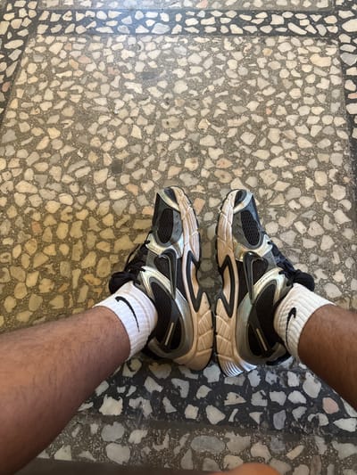 This is a top-down, eye-level, medium shot photograph of a person's legs and feet, wearing white Nike socks with a black Nike logo and black and silver athletic shoes. The person is sitting on a floor made of terrazzo, with a pattern of irregular light-colored stones embedded in a darker gray cement. The floor has a border of darker gray material. The lighting suggests an indoor environment, possibly a home or public space, with natural light casting subtle shadows. The image does not contain any recognizable landmarks or elements that would specifically place it in the city of Dimovo, Bulgaria.