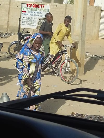 A young girl in a colorful headscarf and patterned dress stands holding a broom on a dusty street. Two boys are nearby, one riding a bicycle and another standing beside him in a dark shirt. In the background is a sign for "Top Quality International School" and a parked motorcycle. The scene appears to be viewed from inside a vehicle, as a dashboard and windshield wipers are visible in the foreground.