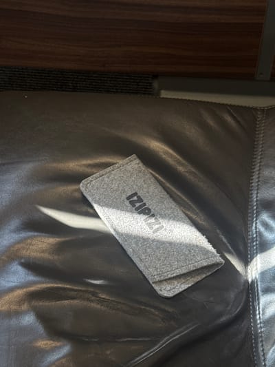 The image shows a close-up view of a gray fabric pouch with black lettering that reads "IZIPPI" placed on a dark, possibly leather, surface. The surface is creased and wrinkled, with a strong shaft of sunlight illuminating it from the upper left, casting shadows and highlighting the texture. To the left of the pouch and dark surface, there is a darker, vertically oriented textured material, possibly a strap or carpet, adjacent to a brown wooden surface with a visible wood grain. The scene is likely indoors, given the artificial lighting implied by the distinct shaft of sunlight, or perhaps near a window. The overall impression is one of a still life, focusing on the texture and interplay of light and shadow on everyday objects. The visible text "IZIPPI" suggests a brand name. No people are visible in the image.