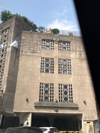 The image shows a building facade in the Hongkou District of China. The building is made of concrete and features a distinctive pattern of square grids with circular openings. The architectural style appears to be from the mid-20th century. The building is partially covered with vegetation, including some trees growing on the upper levels. The photo seems to have been taken from a vehicle, with a portion of the vehicle's interior visible at the top of the frame and a vehicle on the right side of the frame. The sky is visible in the upper left corner.