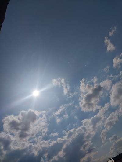 This is a daytime, outdoor, eye-level shot of the sky above Nizhneye Myachkovo, Russia. The sun is prominently visible in the upper left quadrant, emitting bright rays and lens flare. The sky is a clear blue, with scattered white and gray clouds of varying sizes and densities. The clouds appear to be cumulus and stratocumulus formations. In the lower right corner, a portion of a dark-roofed building with a visible chimney is present, suggesting a residential or industrial area. The overall scene depicts a sunny day with a mix of sun and clouds.