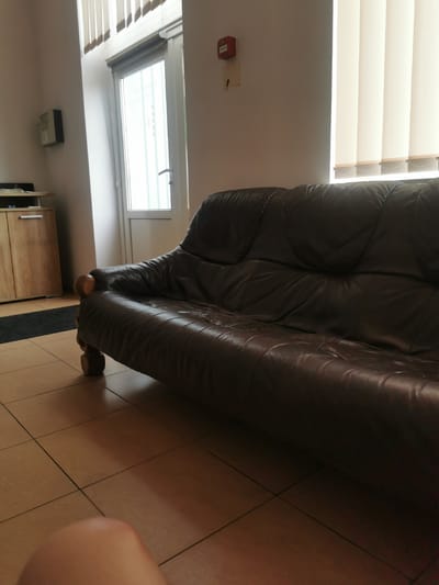 The image shows a brown leather couch in a room. The couch is in the foreground, taking up a significant portion of the frame. To the left of the couch, there is a wooden cabinet with a black rug in front of it. In the background, there is a door with a window and vertical blinds. A red fire alarm is mounted on the wall above the door. The floor is tiled in a light brown color. The scene is dimly lit, with light coming from the window. The image appears to have been taken in a residential or office building. There are no people visible in the image, but a knee is partially visible in the bottom left corner. It is not possible to determine the exact location of the image within Valmiera, Latvia, from the visual information provided.