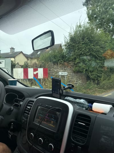 The image is a close-up, interior shot of a vehicle's dashboard and windshield. A smartphone, mounted on a suction cup holder, is visible on the dashboard, displaying a navigation app. To its left, a blue paper cup rests near the windshield. In the background, seen through the windshield, is a red and white striped barrier, and a sign reading "L2630". Beyond the barrier and sign, there is a stone wall covered in dense green foliage. A road with a yellow line is visible to the right. The overall environment appears to be rural, possibly on the outskirts of Bamford Cross, Ireland. The dashboard itself is dark gray and black, with various controls and a touchscreen display visible. A plastic water bottle and a roll of toilet paper are also situated on the dashboard.