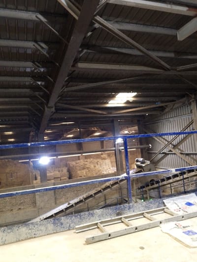 The image shows an interior view of a factory located at latitude 22.693 and longitude 90.601 in Pangasia, Bangladesh. The scene is an industrial area with a ceiling composed of corrugated metal panels. There are two large industrial conveyor belts, one on each side of the image. The conveyor belt on the left side of the image is empty. The conveyor belt on the right side of the image is partially obscured by a large white bag with red and blue lettering. A metal ladder sits on the floor to the right of the conveyor belt. The factory is well-lit by fluorescent lights. The floor is covered with a layer of dust. There are no people or other animals in the image.
