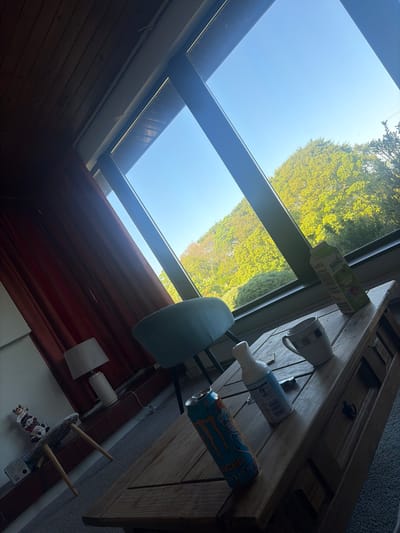 The image depicts an interior scene with a prominent skylight revealing a view of trees and sky, suggesting daylight. A wooden table occupies the upper right portion of the frame, hosting a variety of items including a juice box, a mug, a bottle, and a can of Monster energy drink. A chair is partially visible below the table. Below the skylight is a red curtain and on the floor is a small, round table adorned with what appears to be Christmas-themed figurines and a lamp. The image provides no clear indicators of a specific location within The Municipal District of Adare — Rathkeale, Ireland, only an interior space.