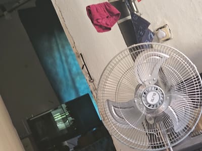 The image depicts an indoor scene with a standing fan in the foreground, its metallic blades and grill clearly visible. Above and behind the fan, a pair of red undergarments with lace trim are hanging from a thin wire. To the left of the fan, a dark-colored monitor screen displays a green, abstract pattern. The background consists of a textured wall and a blue-colored section, possibly a curtain or a painted surface, with light reflecting on it. A small, white object, possibly a light fixture or a sensor, is mounted on the wall to the right. The overall impression is that of a domestic living space. The image does not contain any information that would allow for its specific location within Daskotna, Bulgaria.