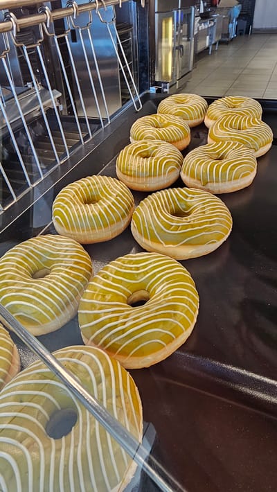 A close-up, high-angle shot captures a display of approximately ten green-frosted donuts with white drizzles. The donuts are arranged on a black tray, with some in focus and others slightly blurred. Behind the donuts, a metal rack with horizontal bars and vertical supports is visible, suggesting a kitchen or bakery setting. The background is out of focus, showing what appears to be commercial kitchen equipment, including stainless steel surfaces and cabinetry. The lighting is bright, casting reflections on the glossy frosting of the donuts and the metal elements. No people are visible in the image, and the scene appears to be a static product display. This scene could be located within a bakery or cafe in Cuarte de Huerva, Spain, showcasing freshly prepared pastries for sale.
