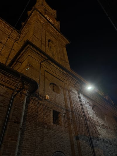 The image shows the bell tower of a brick church at night in Lagnasco, Italy. The bell tower is illuminated by artificial lights, casting shadows on the brickwork. A clock face is visible near the top of the tower. The surrounding sky is dark. The image is taken from a low angle, looking up at the tower. There are no people or animals visible in the image.