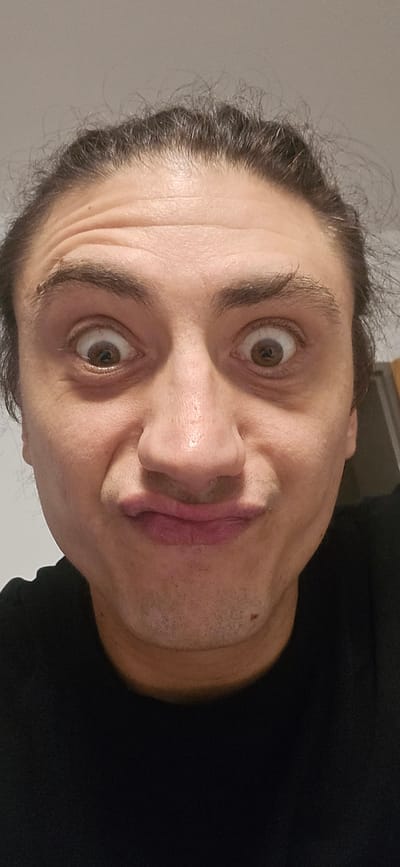 This is a close-up, front-facing selfie of a person with dark hair pulled back. The person is making a comical expression with wide eyes and pursed lips. Their forehead displays noticeable wrinkles. The background is a plain, light-colored wall. The image was captured indoors, likely in a residential setting. There is no discernible information about the location in Bad Fallingbostel, Germany.