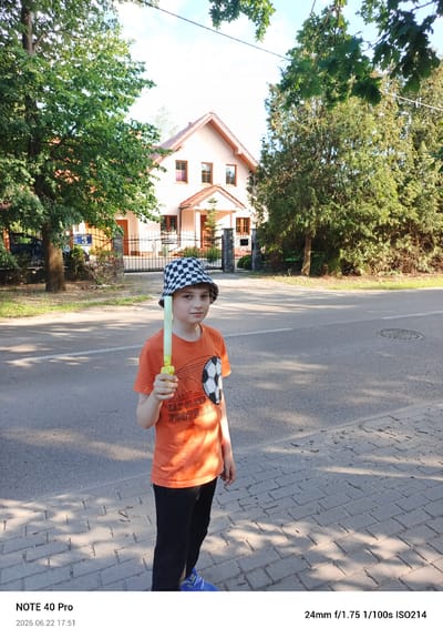 A young boy wearing an orange t-shirt with a soccer ball graphic and black pants stands on a sidewalk. He is also wearing a black and white checkered bucket hat and blue sneakers. He holds a glowing yellow toy in his right hand. The t-shirt has the text "EAT SLEEP FOOTBALL" printed on it.

The setting appears to be a suburban street in Kamionka, Poland. A light-colored house with a red tile roof is visible behind him, along with trees and a metal fence. The boy is standing on a brick sidewalk next to a paved road. Sunlight casts shadows on the ground, suggesting it is daytime. The image is dated 2026.06.22 at 17:51, indicating it was taken in the late afternoon or early evening. The weather appears to be clear and sunny. The boy has a neutral expression.