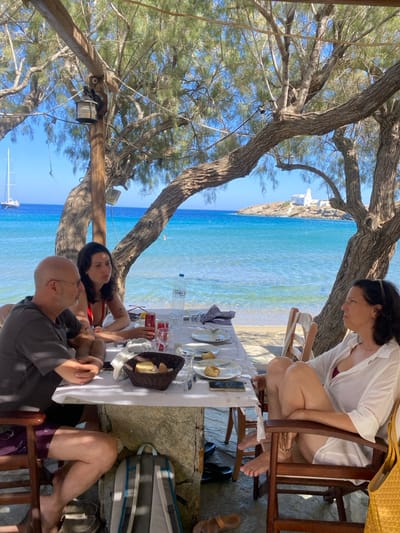 The image depicts an outdoor dining scene at a seaside establishment in Faros, Greece. Three individuals are seated at a white-tablecloth-covered rectangular table positioned on a sandy surface directly adjacent to the sea.
To the left, a bald, middle-aged male wearing glasses, a dark gray t-shirt, and dark shorts is seated, with his hands clasped on the table. His dark sandals are visible on the ground. Opposite him, a younger female with dark hair, wearing a red bikini top, faces away from the camera, looking towards the right. To the right, a middle-aged female with dark hair, wearing a white button-down shirt over a red top, is seated, looking towards the bald male. Her legs are crossed, and a yellow woven bag rests beside her chair.
On the table are multiple white plates, some containing food remnants, several empty glasses, a dark woven basket with bread, a clear plastic water bottle (Zagori brand), two red Coca-Cola cans, and a black smartphone. A striped napkin is also present. Under the table, a gray and blue backpack is visible next to a pair of dark sandals.
The dining area is shaded by the natural canopy of large trees with gnarled trunks and green foliage. Wooden beams and a hanging lantern are visible above. The background features calm, clear turquoise-blue water meeting a sandy beach. In the mid-distance, a distinct white-washed building complex is situated on a rocky islet or promontory. Further to the left in the bay, a large white two-masted sailboat is anchored. The scene is illuminated by bright daylight, casting strong shadows.
