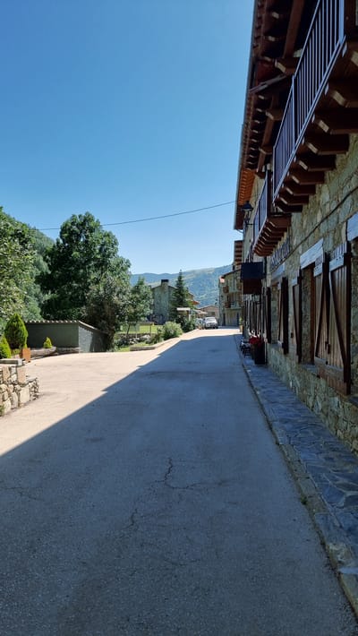 The image depicts a narrow, paved street in a village setting, likely Setcases, Spain. The perspective is rotated 90 degrees clockwise, making the scene appear on its side. On the left side of the street, a traditional stone building features wooden shutters and balconies, suggesting rustic architecture common in mountainous regions. The building's facade is made of rough-hewn stones, with wooden beams and elements adding to its charm.

The street itself is asphalted and appears well-maintained, with a shadow cast across it from the right, indicating it is daytime. On the right side of the street, there is a glimpse of a green grassy area with some vegetation and stone elements, hinting at a yard or garden. Further down the street, more buildings are visible, interspersed with trees and hills in the background, suggesting a rural or semi-rural location within a valley or mountainous terrain.

A white van is parked further down the street, and a few other structures blend into the background. The sky is a clear, bright blue, indicating fair weather and a sunny day. The overall impression is one of a quiet, picturesque village, bathed in sunlight. No specific activity or event is clearly visible, but the scene suggests a typical day in a European village. There is no visible text in the image.