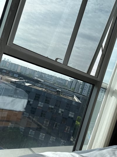 The image shows a view through a slanted window of buildings in Minhang District, China. The window has a screen and is partially open. The buildings are a mix of residential and commercial structures, with some featuring dark facades and many windows. The sky is cloudy. A white curtain is visible on the right side of the frame. The perspective suggests the photograph was taken from a higher floor.
