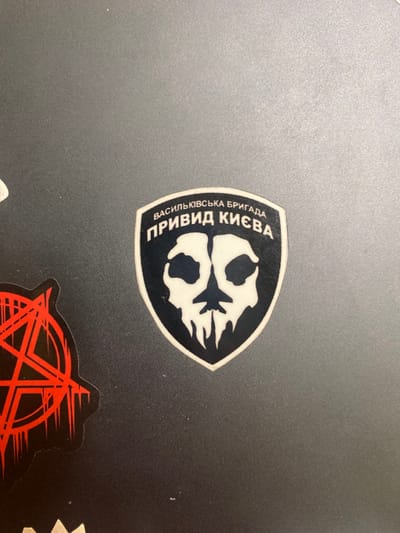 The image shows a sticker on a dark grey surface. The sticker is a black shield with a white skull inside. The text "Васильківська бригада" (Vasylkivska Brigade) and "ПРИВИД КИЄВА" (Ghost of Kyiv) is written in white on the sticker. The coordinates 37.574, -77.541 correspond to a location in Richmond, Virginia, United States. There are no people or subjects in the image. The surface appears to be a laptop. There is a second sticker, an anarchy symbol in black and red, visible in the lower left hand corner of the image. The anarchy symbol is partially obscured by the shield sticker.