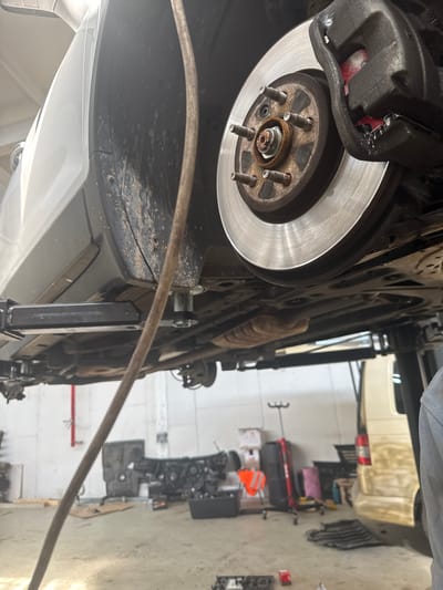 A car's brake rotor and caliper are visible, suspended on a car lift in a workshop. The car is white and appears to be a passenger vehicle. The workshop floor is concrete and shows some dirt and debris. In the background, other vehicles, possibly vans, and various workshop equipment, including an engine, are present. There are no people clearly visible in the frame. The scene suggests ongoing automotive repair work. The location is stated to be in Schkopau, Germany.