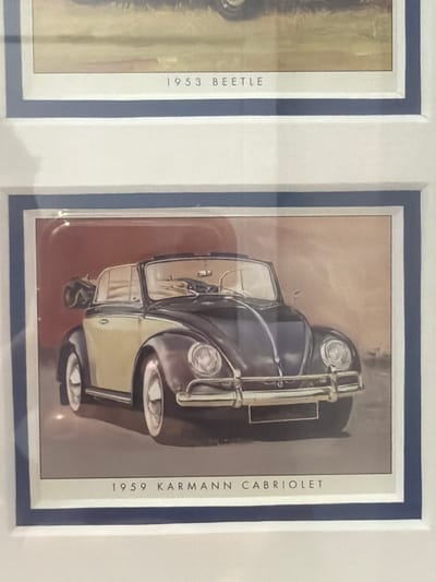 This is a photograph of two framed prints of Volkswagen cars. The upper print depicts a 1953 Beetle. The lower print shows a 1959 Karmann Cabriolet, a convertible version of the Beetle, in a two-tone color scheme of dark grey and tan. The cars are illustrated in a realistic style. The image does not contain any information about the city of Ouahat Sidi Brahim, Morocco.