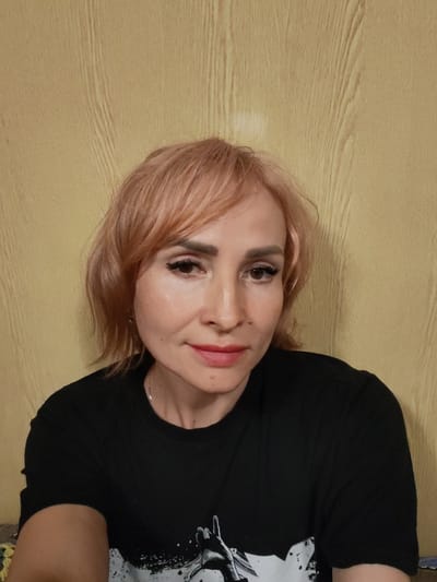 The media file is a close-up portrait of a woman with light pink, short hair. She is wearing a black t-shirt with a graphic print that is partially visible. Her makeup includes eyeliner, mascara, and lipstick. She has a light skin tone and appears to be looking directly at the camera with a neutral expression. The background is a yellow wall with a wood grain texture. There is no visible text in the image. The lighting suggests an indoor setting, possibly during the day, but no specific time or weather can be determined. The location context of Annensky Most, Russia, is provided but not directly observable from the image itself.