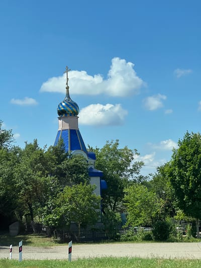 An Orthodox church, characterized by a blue roof and a gold-and-blue striped dome topped with a cross, is partially obscured by lush green trees. The church is situated in Novomyshastovskaya, Russia. The sky above is a clear blue with scattered white clouds. In the foreground, there is a gravel road with white posts featuring red reflectors, bordered by grass. A black metal fence runs along the property line, separating the road from the church grounds.