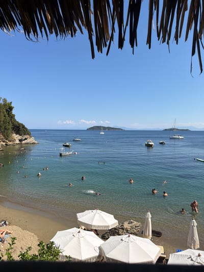 A sun-drenched beach scene unfolds in Skiathos, Greece. The image captures a vibrant day with people enjoying the clear, blue water and sandy shore. Several individuals are swimming and wading in the shallow sea, with some further out in deeper water. White beach umbrellas are strategically placed along the sand, offering shade.
In the background, various boats are anchored or cruising in the tranquil waters of a bay. A sailboat with a tall mast is prominent, alongside smaller motorboats. The coastline is visible, featuring rocky outcrops and lush green vegetation, characteristic of a Mediterranean landscape. The sky is a brilliant, cloudless blue, indicating fair weather and likely midday. The overall atmosphere is one of relaxation and leisure, with people embracing the summer warmth and the beauty of the Greek coast. The foreground shows a thatched-like structure, possibly part of a beach umbrella or canopy, framing the view.