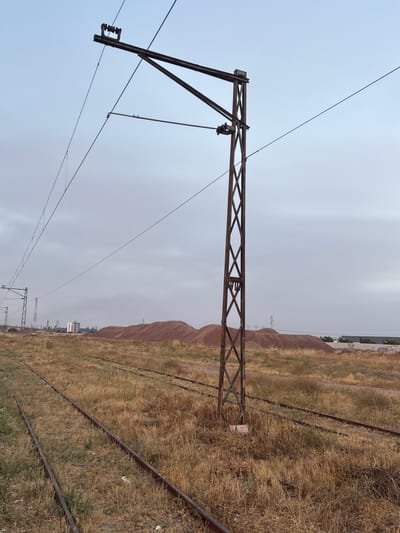 The image depicts an outdoor scene in Berrechid, Morocco, characterized by an abandoned industrial or railway area. In the foreground, multiple sets of disused railway tracks cut through dry, overgrown grass. A prominent, rust-covered metal pylon supporting overhead electrical wires stands to the right of the frame, its lattice structure visible against a pale, overcast sky. In the midground, large mounds of reddish-brown material, possibly gravel or earth, are piled up. In the distance, several industrial buildings and structures, including a multi-story building and utility poles, are visible, suggesting proximity to an active industrial zone. The overall impression is one of disuse and decay within a potentially industrial landscape. There are no people or animals present in the image, and no actions or interactions are occurring.