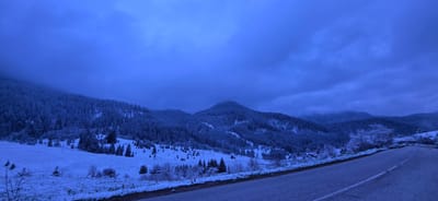 This wide-angle landscape photograph captures a serene, snow-covered mountain scene in Stoikite, Bulgaria, bathed in a profound blue hue. The pervasive blue tint suggests either twilight (dawn or dusk) or an extremely dark, overcast winter day, contributing to a tranquil and slightly melancholic atmosphere.

In the foreground, a dark, paved road with a faint white dividing line curves gently from the lower right towards the center-right of the frame, leading the eye into the distance. To the left of the road, a vast, undulating field or valley is blanketed in fresh, undisturbed snow. Scattered individual coniferous trees, their branches laden with snow, dot this open area, alongside some bare deciduous trees and shrubs that appear frosty.

The midground and background are dominated by a series of snow-dusted mountains and hills, densely covered with coniferous forests. The trees are heavily caked in snow, outlining their dark forms against the lighter snowy slopes. A prominent, conical peak rises in the center-left midground, also covered in forest and snow, with hints of mist or low clouds obscuring its upper reaches. Patches of fog or heavy cloud cover also cling to the higher elevations and distant valleys, adding to the atmospheric depth.

The sky is a uniform, heavy dark blue, filled with thick, textured clouds that extend across the entire upper half of the image, allowing very little direct light to penetrate. The overall weather condition is wintery and cold, with a fresh blanket of snow. There are no people, animals, vehicles, or discernible man-made structures beyond the road visible, emphasizing the raw, untamed beauty and quiet isolation of the winter wilderness. No visible text is present in the image.