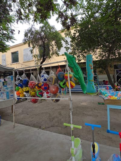In the city of Syrdarya, Uzbekistan, a street market stall displays an array of children's toys. Hanging from a metal rack are numerous inflatable balls, some designed to resemble soccer balls, others with colorful patterns. To the right of the balls, an inflatable green crocodile toy is suspended, followed by a blue inflatable punching bag decorated with cartoon figures. In the foreground, three children's scooters are visible, two of which are partially wrapped in plastic. Behind the stall, a light yellow building with boarded-up windows stands, suggesting ongoing construction or renovation. Trees with green foliage partially obscure the building. The ground is a mix of concrete and dirt, with a small blue table in the background holding a box of water guns and other toys. The scene is brightly lit by natural daylight.