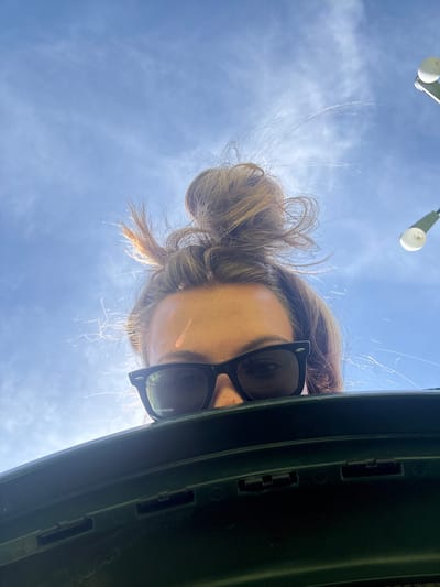 An upward-facing shot features a person wearing sunglasses with their hair in a messy bun. They are looking down at the camera, and the bright sky is visible behind them. A green object is on the right. Two white lights are partially visible in the top left corner against the clear blue sky.