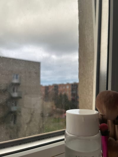The image depicts a view through a dirty window. Outside, the sky is overcast. A blurred building is visible in the background, suggesting an urban setting, possibly Raduga, Russia, as requested. In the foreground, resting on the window sill, are several makeup brushes and a cylindrical white bottle, partially obscured. The scene appears still, with no apparent human activity. The overall impression is one of neglect, as evidenced by the dirty window.