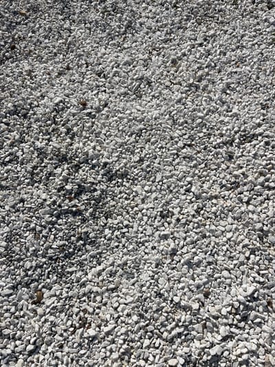 The image shows a ground covered in white gravel. The gravel is densely packed, with minimal space between the stones. The gravel is a mix of small and medium-sized white stones, with some darker grey stones interspersed throughout. The gravel is illuminated by sunlight, which highlights the texture of the stones and creates a sense of depth. The image was taken in Limenaria, Greece, at latitude 40.626, longitude 24.57. The image does not contain any people or objects.