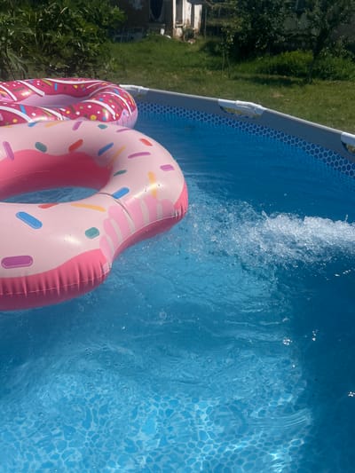 In Belo Pole, Bulgaria, an above-ground swimming pool is filled with clear blue water. Two inflatable donut-shaped pool floats, one pink with colorful sprinkles and another pink with white icing and red accents, are floating on the surface. The pool is surrounded by green grass and trees, with a rustic building visible in the background under a bright sky. The water shows signs of movement, with splashes and ripples indicating a potential water jet or disturbance.