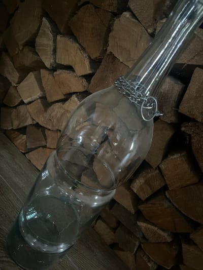 A clear glass bottle with a chain wrapped around its neck lies diagonally on a wooden surface. The bottle has a bulbous body and a narrow neck. The background is a wall of stacked, cut firewood. The lighting is dim, casting shadows and highlighting the textures of the wood and glass. There are no people present in the image.